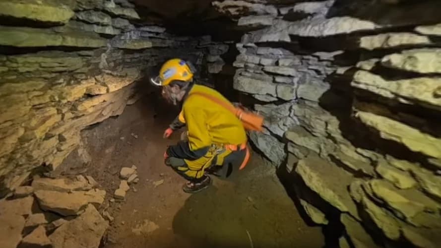 Spéléologue accroupi dans une galerie basse de la Caverne de Saint-Léonard, casque et combinaison